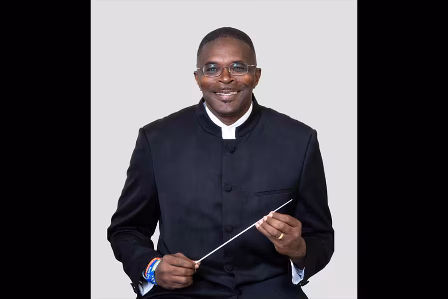 Portrait of conductor Shelby Chipman smiling while seated against a light gray background, wearing glasses and a black formal jacket, holding a white conducting baton in both hands.