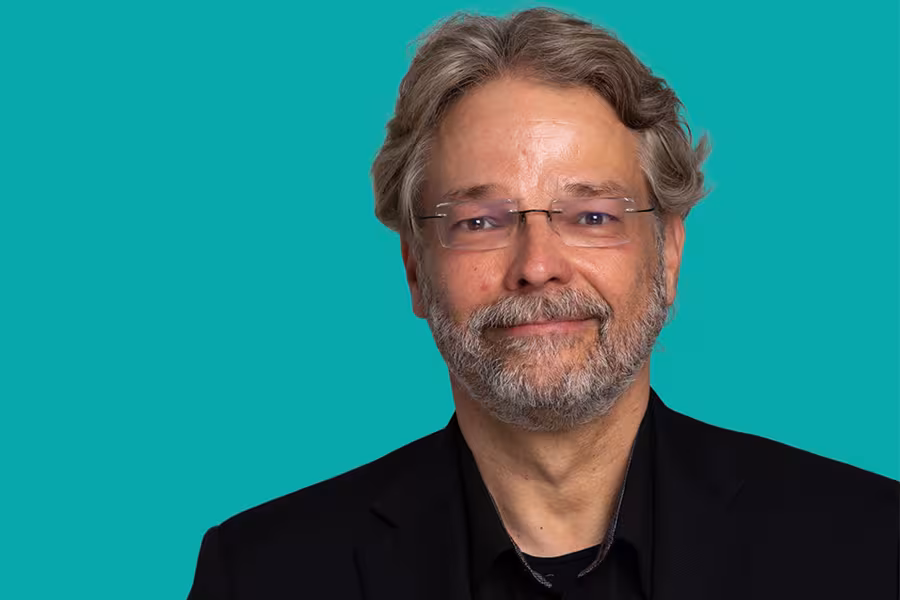 Portrait of a smiling man with gray hair, beard, and glasses wearing a dark jacket and black shirt against a teal background.