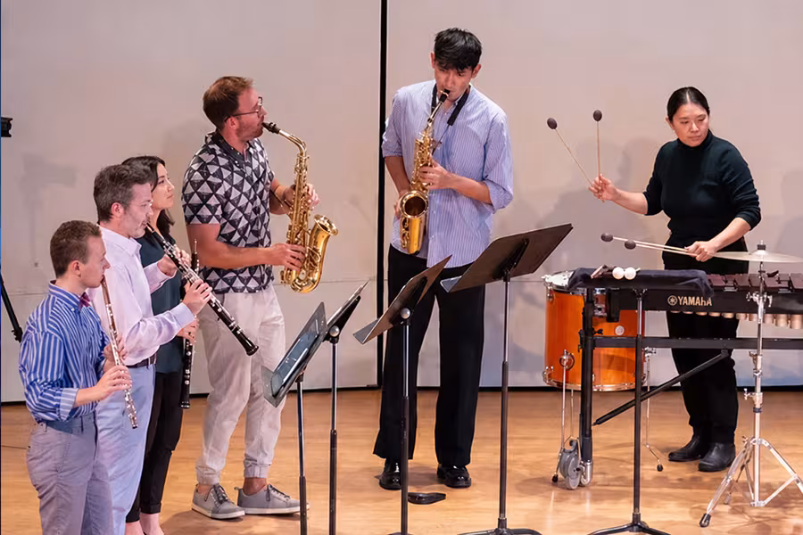A group of musicians perform together onstage during a chamber music concert. Three woodwind players and two saxophonists stand beside a percussionist playing a mallet instrument, surrounded by music stands under warm stage lighting.