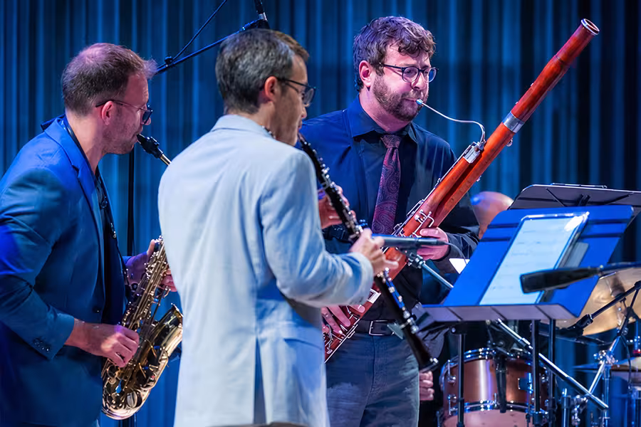 Three musicians perform on stage under blue concert lighting during a live woodwind ensemble performance. A saxophonist, clarinetist, and bassoonist stand closely together beside music stands and percussion instruments, creating an intimate and energetic chamber music atmosphere.