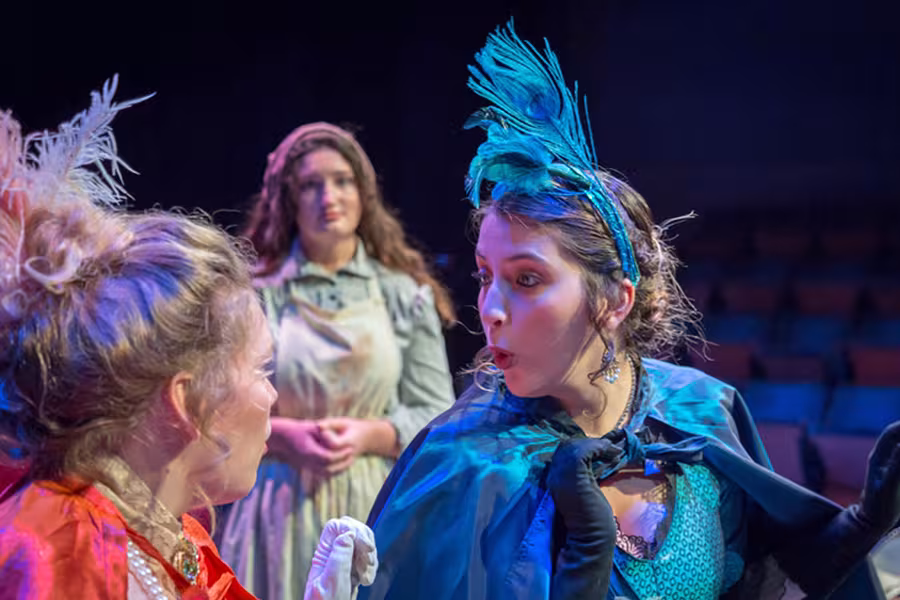 Students perform an expressive scene during a theatrical production, wearing colorful period-inspired costumes and feathered headpieces under blue and purple stage lighting in an auditorium.