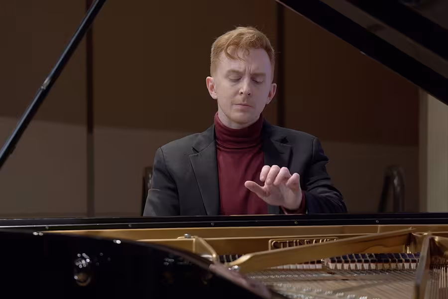 A Chris Goodpasture with short red hair performs at a grand piano onstage, wearing a black blazer over a burgundy turtleneck. His eyes are closed in concentration as one hand hovers above the keys. The open piano lid and strings are visible in the foreground, with warm wood paneling in the background.