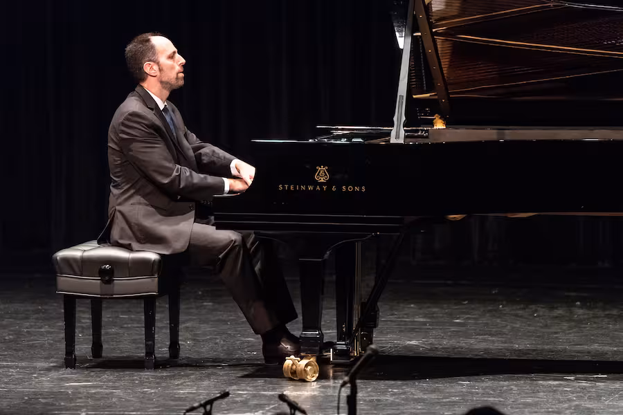 Spencer Myer sits at a black Steinway & Sons grand piano on a dimly lit stage, performing in a dark suit and tie. He is shown in profile, focused on the keyboard while seated on a black piano bench, with the open lid of the grand piano extending across the stage.