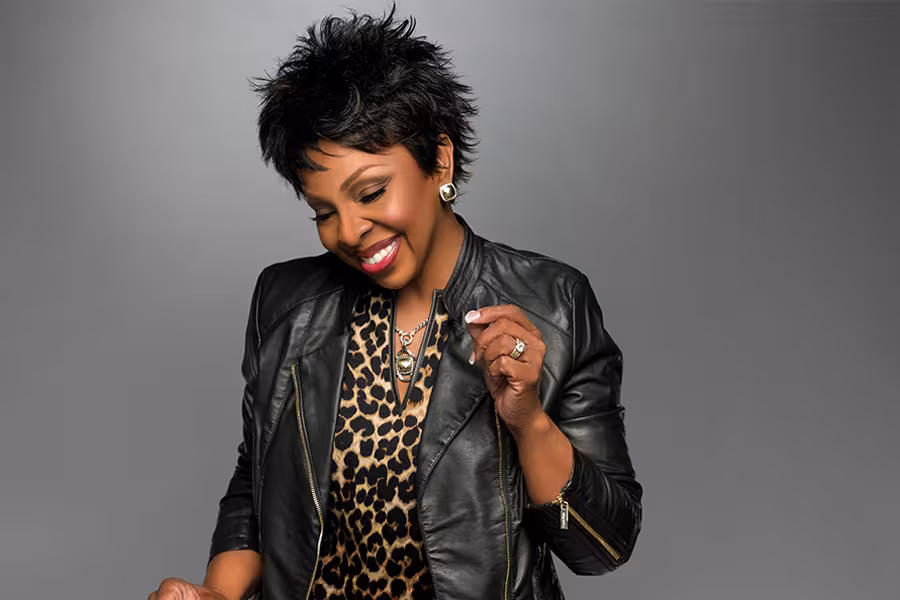 Gladys Knight smiles warmly while looking down, wearing a black leather jacket over a leopard print top, with short styled hair and gold jewelry, posed against a neutral background.