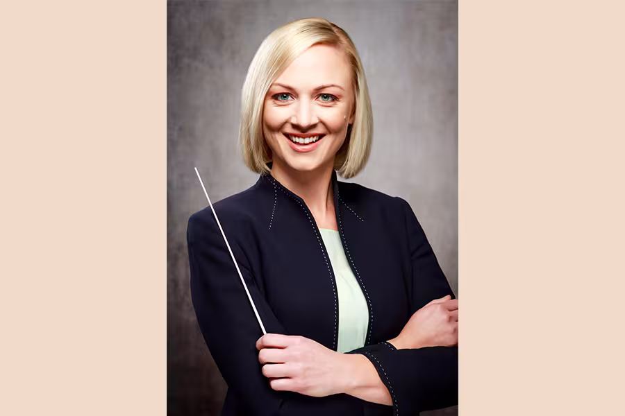 Professional portrait of a smiling blonde Kara Huber holding a conductor’s baton while standing with her arms crossed. She is wearing a dark blazer over a light blouse against a neutral gray studio background.