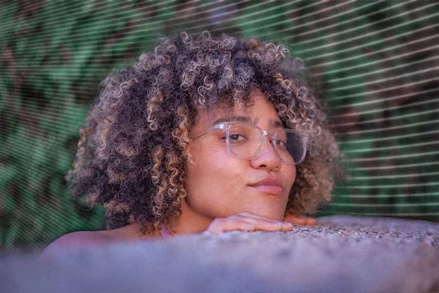 Portrait of poet Sasha Debevec-McKenney resting her chin on folded hands, looking toward the camera with a calm expression. She has curly highlighted hair and wears large clear-framed glasses. The background features soft green foliage and diagonal lines, creating a dreamy, artistic effect.