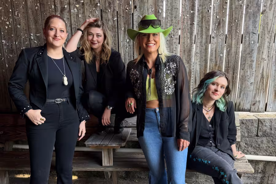Four musicians pose together in front of a rustic wooden wall. One woman in a bright green cowboy hat and sequined black shirt stands smiling in the center, while the other three, dressed mostly in black, are positioned around a wooden bench. One seated musician has teal-colored hair, and the group appears relaxed and casual outdoors.