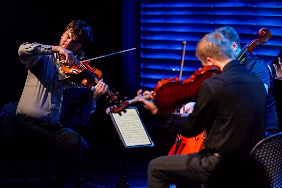 Members of Viridian Strings perform a chamber music concert on stage under blue lighting. In the foreground, string players are seated in a semicircle, focused on their instruments and sheet music. A violinist at left plays with expressive intensity while fellow musicians perform nearby, creating an intimate and engaging concert atmosphere.