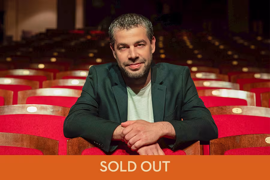 Portrait of the Jader Bignamini, conductor, seated among red theater seats with the words "SOLD OUT" displayed across the bottom of the image.