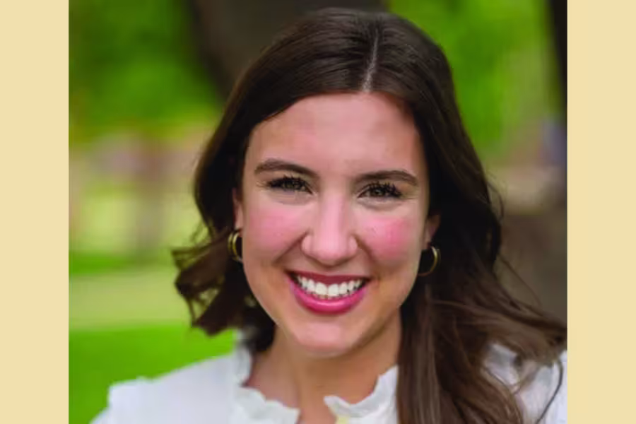 Head-and-shoulders portrait of a smiling Grace Jackson with shoulder-length dark brown hair, wearing gold hoop earrings and a white blouse with a ruffled neckline. The background is softly blurred with green trees and lawn, creating an outdoor setting.