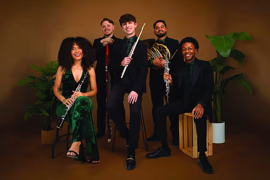 Five members of the ZaRiah wind quintet pose together in a studio with warm brown backdrop and green plants. From left to right are a clarinetist seated in a green velvet outfit, a bassoonist standing behind, a flutist seated in the center, a French horn player standing behind, and an oboist seated on a wooden box. All wear coordinated black and green concert attire and hold their instruments while smiling toward the camera.