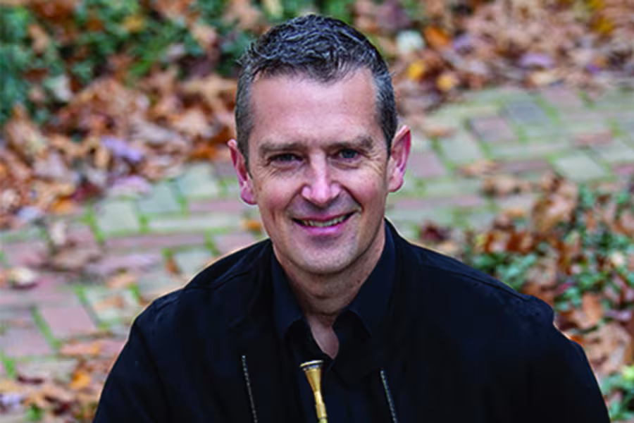 Portrait of horn player Adam Unsworth smiling at the camera outdoors, wearing a black shirt with his French horn visible at his chest.