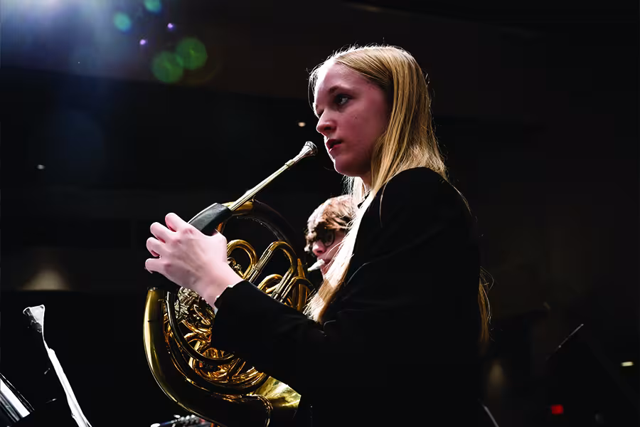 Student musician performing on a French horn during a concert rehearsal, holding the instrument with focused concentration while fellow ensemble members appear softly blurred in the background.