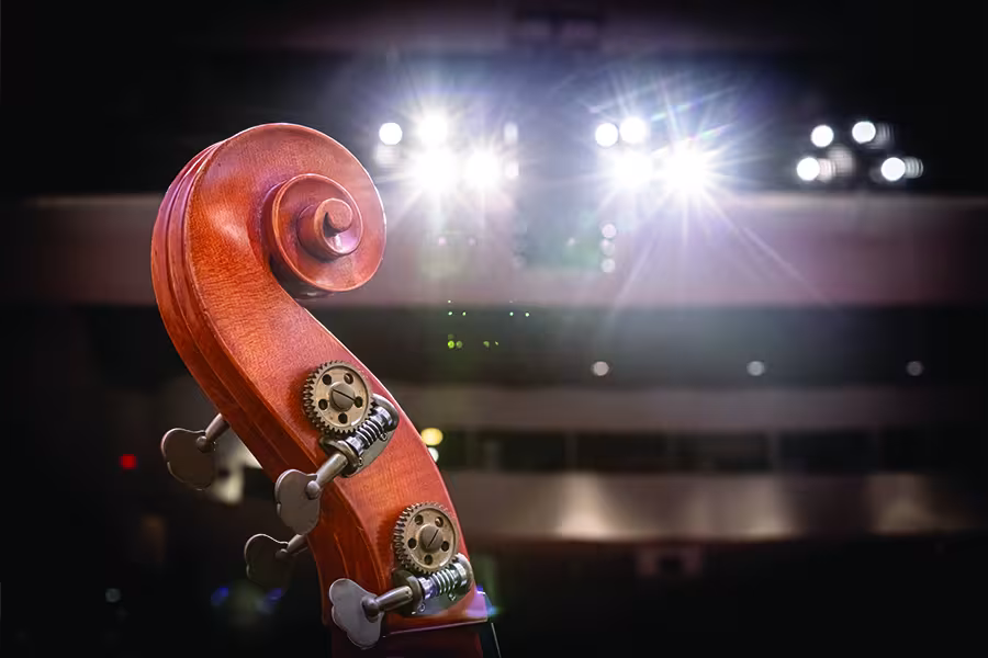 Close-up of the scroll and tuning pegs of a string instrument, illuminated by bright stage lights in a concert hall with the audience seating softly blurred in the background.