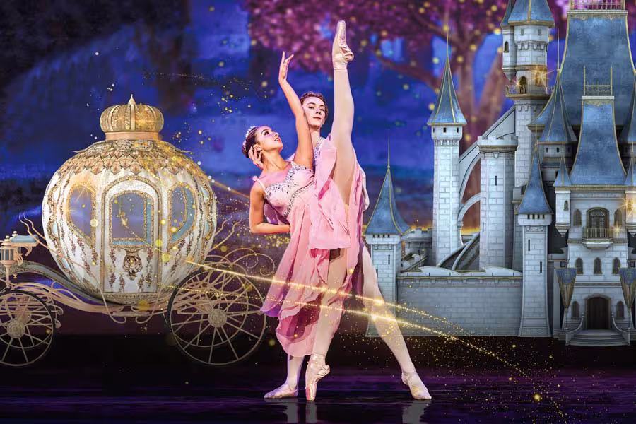 Two ballet dancers performing on stage, with one dancer in a pink costume extending her leg upward and another dancer standing behind her. A dark, moonlit forest scene appears in the background.