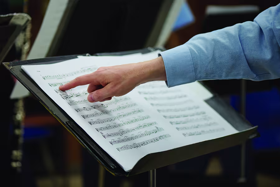Close-up of a hand pointing to sheet music on a music stand during a rehearsal, with instrument stands and musicians softly blurred in the background.
