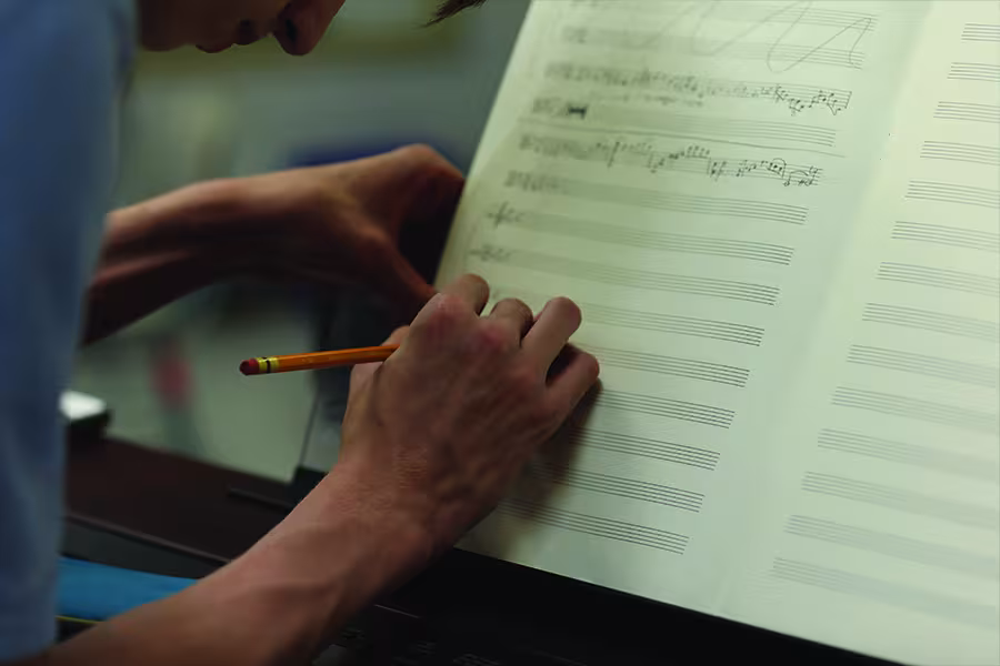 Close-up of a composer writing musical notation with a pencil on a score, with handwritten notes and blank staff lines visible on the manuscript paper.