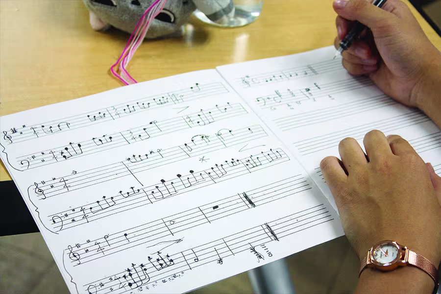 Close-up of a student composing music by writing notation on sheet music with a pen, with completed measures and blank staff lines visible on the score.