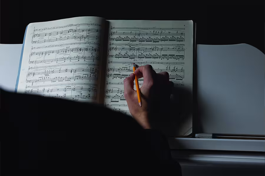 Close-up of a musician marking notes with a pencil in an open musical score on a piano music stand, illuminated by focused light in a quiet practice setting.