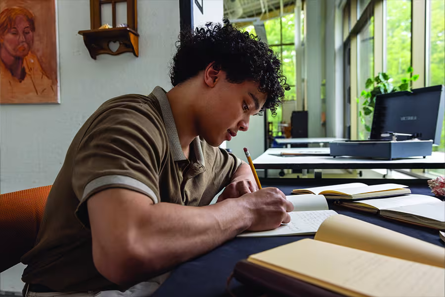 Student sits at a table writing in a notebook with a pencil, surrounded by open journals in a bright studio classroom with large windows and artwork in the background.