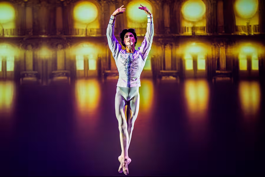 A ballet dancer in a white costume performs on stage with arms raised overhead, set against a warmly lit, ornate interior backdrop with glowing golden windows and reflections on the floor.