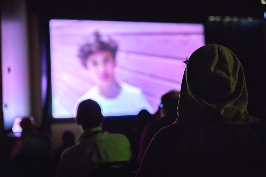 Audience members watch a film screening in a dark theater as a movie plays on a large projection screen, with viewers silhouetted against the light from the screen.