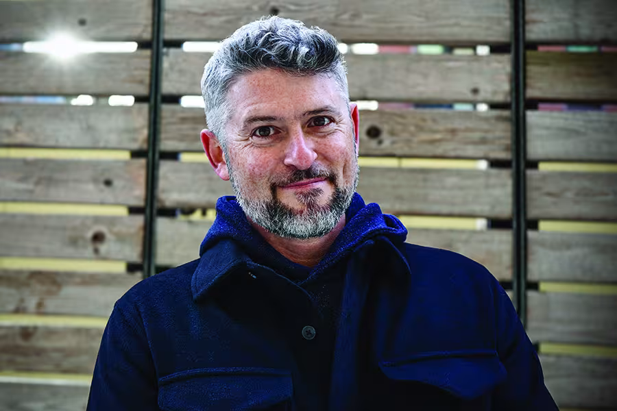 Martin Rock, a middle-aged man with short gray hair and a gray beard, smiling at the camera while wearing a dark jacket, photographed against a wood-paneled background.