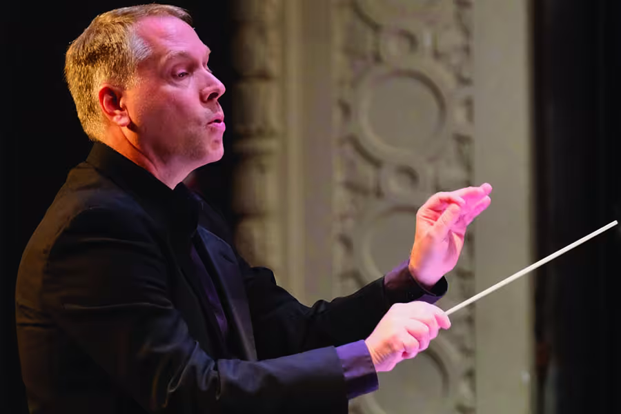 Conductor Morehouse conducts an orchestra, holding a baton in his right hand and raising his left hand to guide the musicians.