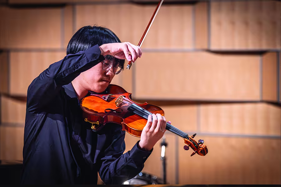 Student violinist performs during a recital, playing the violin with focused expression while holding the bow above the strings on a concert hall stage.