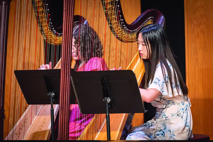 Two student harpists perform together on stage, each playing a concert harp behind music stands during a chamber music recital.