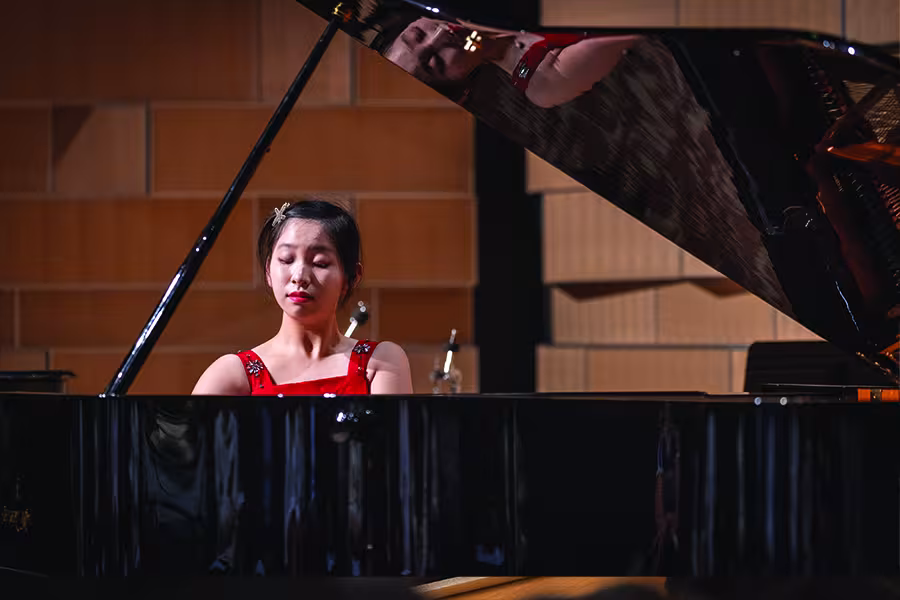 Student pianist performs at a grand piano during a recital, wearing a red concert dress, with her reflection visible in the raised piano lid on a concert hall stage.
