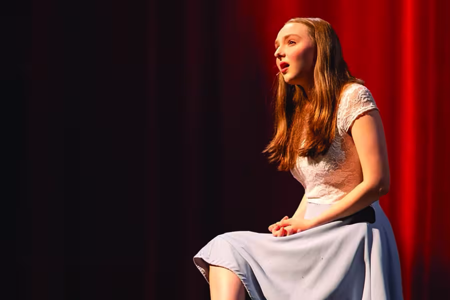 Young woman singing on stage, seated against a dark background with red stage lighting.