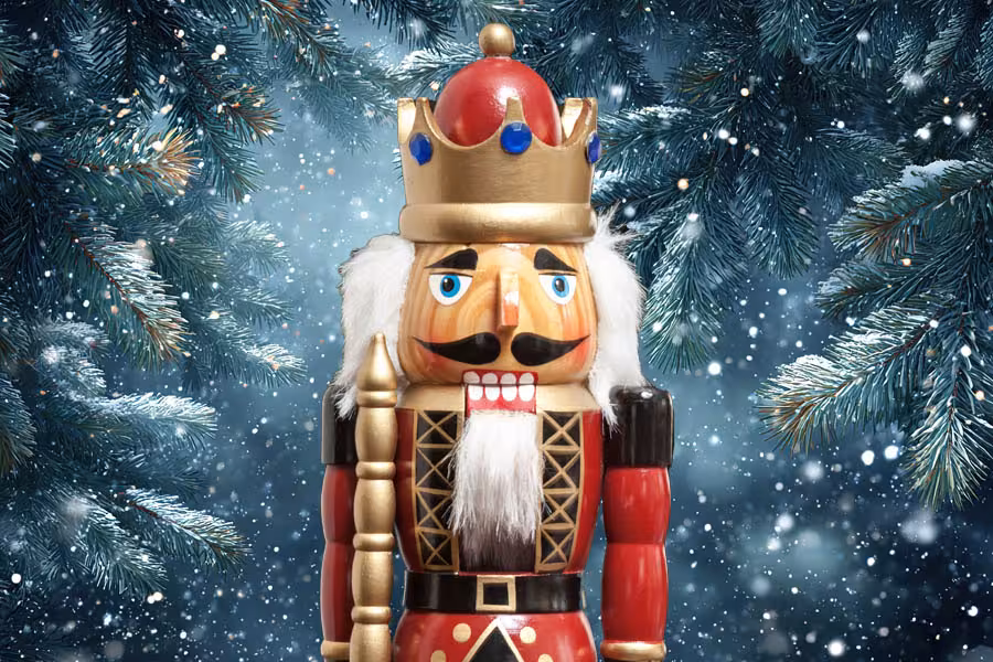 A colorful wooden nutcracker wearing a crown stands among snow-covered evergreen branches, surrounded by falling snow.