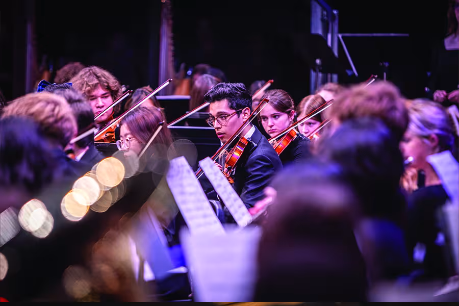 Student orchestra performs on stage, with violinists playing together while surrounded by other musicians and music stands.