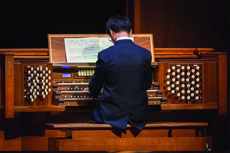 Student organist performs at a large pipe organ, seated at the console with sheet music open on the music stand.