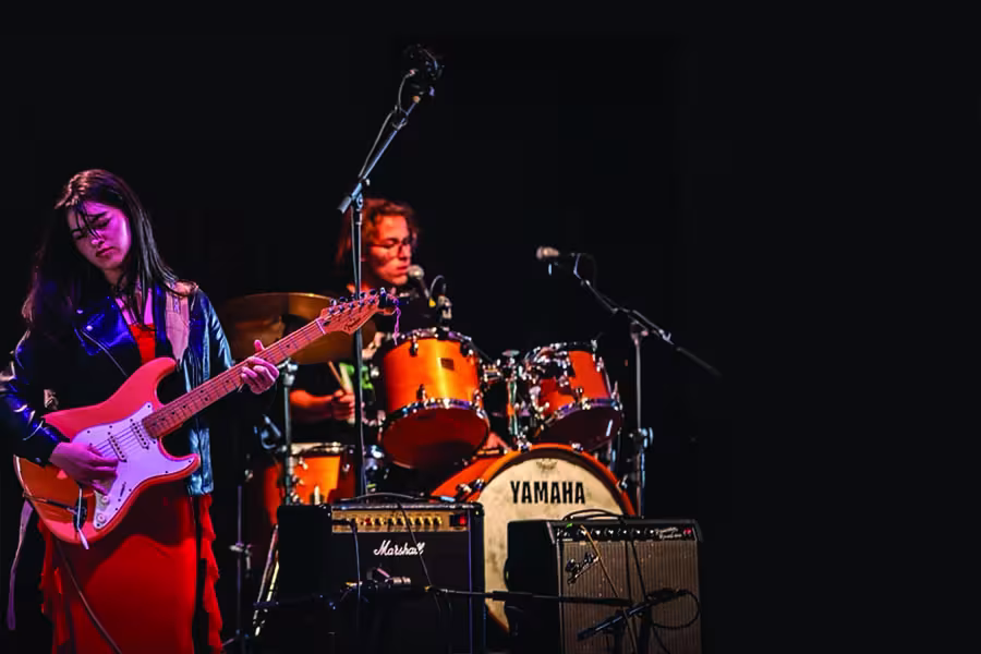 Musician playing electric guitar with a drummer performing behind her on stage.