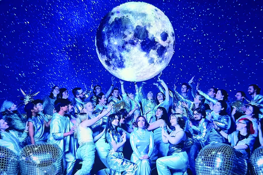 A group of performers in shimmering silver costumes gather beneath a large glowing moon, reaching toward it amid a star-filled blue backdrop.