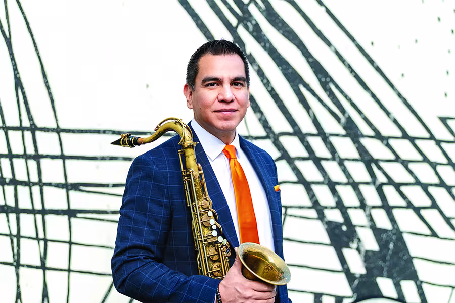 A man in a blue suit and orange tie poses with a saxophone in front of a black-and-white abstract mural.