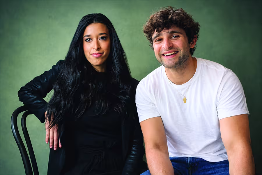 Portrait of two poets seated side by side, a woman with long dark hair wearing black and a man with curly brown hair wearing a white shirt, both smiling at the camera.