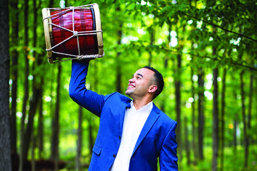 Percussionist, Jean Carlo Urena Gonzalez, in a blue suit raises a large red drum overhead while smiling in a sunlit forest.