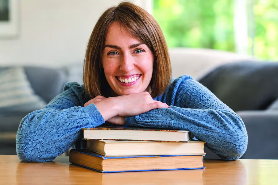Jillian Laine smiles at the camera while resting her arms on a stack of three books at a table.