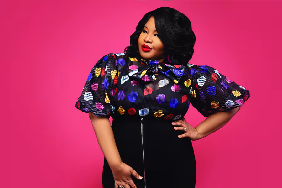 Portrait of a woman with black hair wearing a colorful patterned blouse and black skirt against a bright pink background.