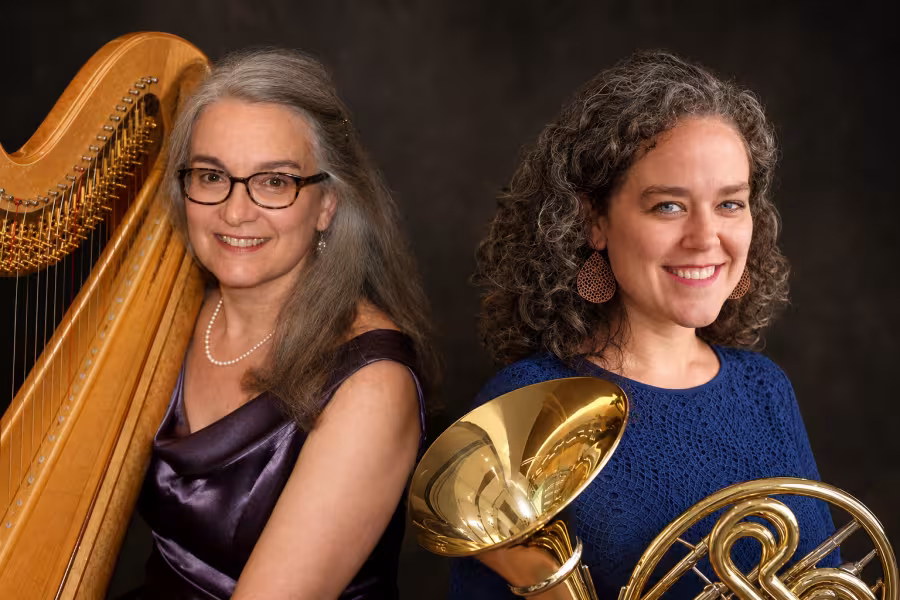 Two smiling musicians pose with their instruments against a dark background. A harpist stands beside a large harp, while a horn player holds a French horn.