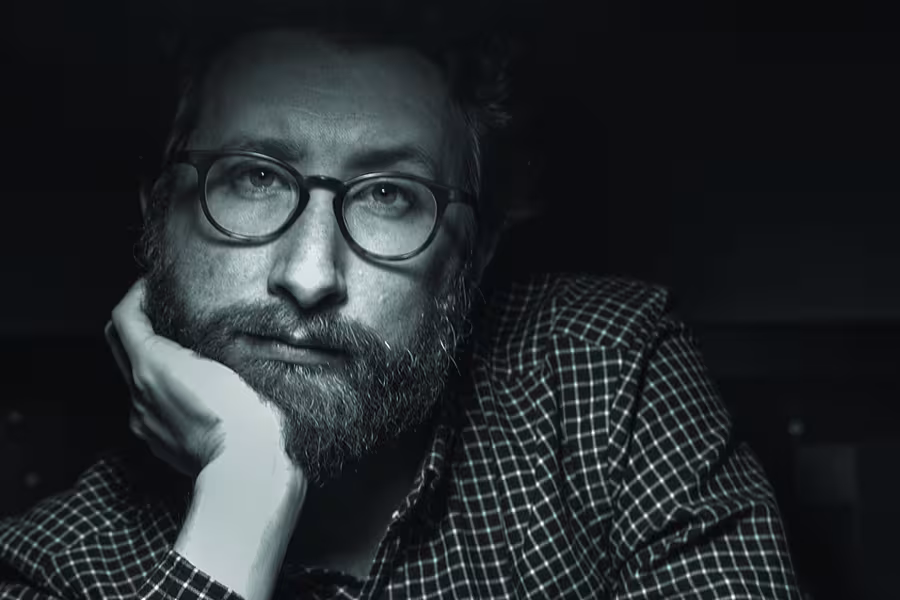 Black-and-white portrait of poet Malachi Black wearing glasses and a plaid shirt, resting his face on his hand and looking toward the camera.
