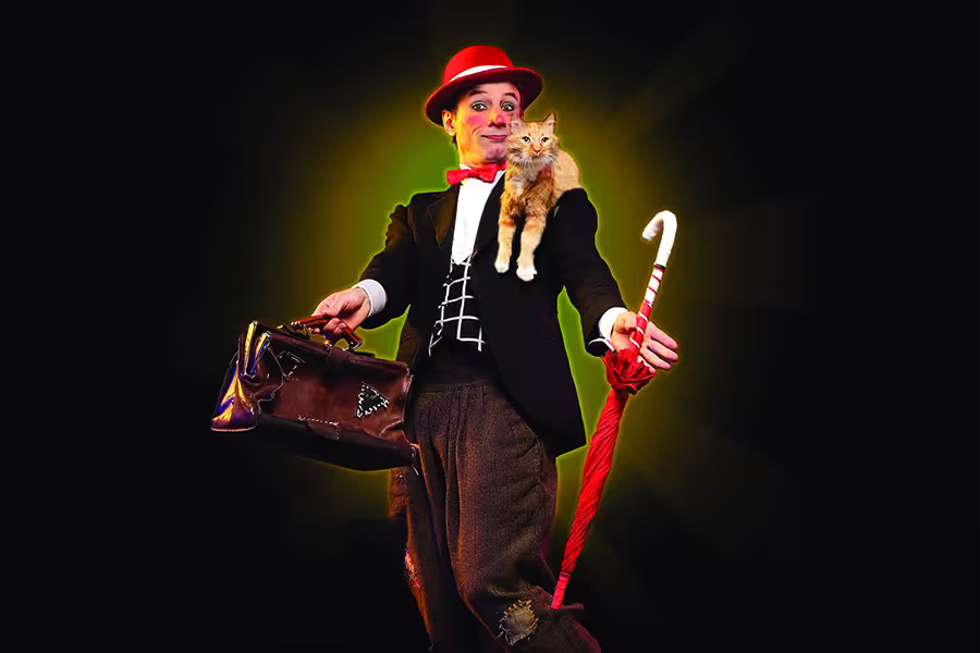 Performer in a red hat and bow tie holds an orange cat and a red umbrella, with a brown suitcase in the other hand, against a black background.