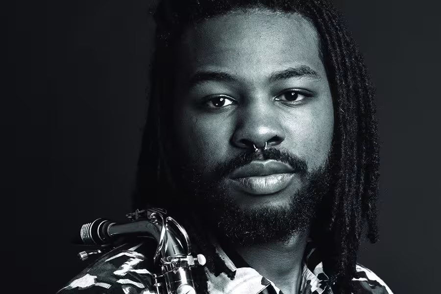 Black-and-white portrait of Kameron Sheffield with long locs and a beard, wearing a patterned shirt and holding a saxophone over his shoulder against a dark background. with long locs and a beard, wearing a patterned shirt and holding a saxophone over his shoulder against a dark background.