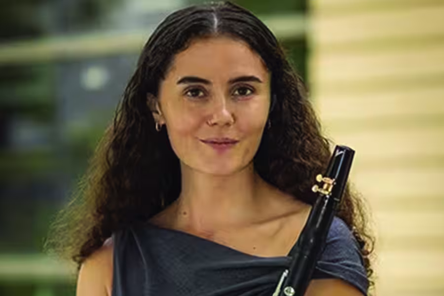 Portrait of Katia Sofia Waxman with long, curly dark hair wearing a dark blue top and holding a black woodwind instrument against a softly blurred indoor background.