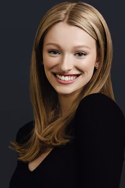 Headshot of a smiling young woman with long blonde hair and blue eyes. She is wearing a black top and small stud earrings and is turned slightly toward the camera. The portrait is professionally lit against a dark blue-gray studio background.