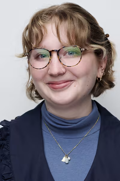 Headshot of a smiling young woman with short blonde hair and round tortoiseshell glasses. She is wearing a blue turtleneck layered under a navy top, along with several earrings, a nose ring, and a pendant necklace. The portrait is set against a light gray studio background and is softly lit.