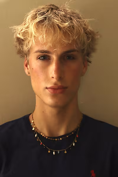 Headshot of a young man with tousled blond hair looking directly at the camera with a neutral expression. He is wearing a dark blue shirt, small stud earrings, and layered necklaces. Warm natural light illuminates his face against a simple beige background.